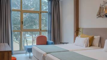 A hotel room with a large window, a bed, a chair, a small table, and a lamp.