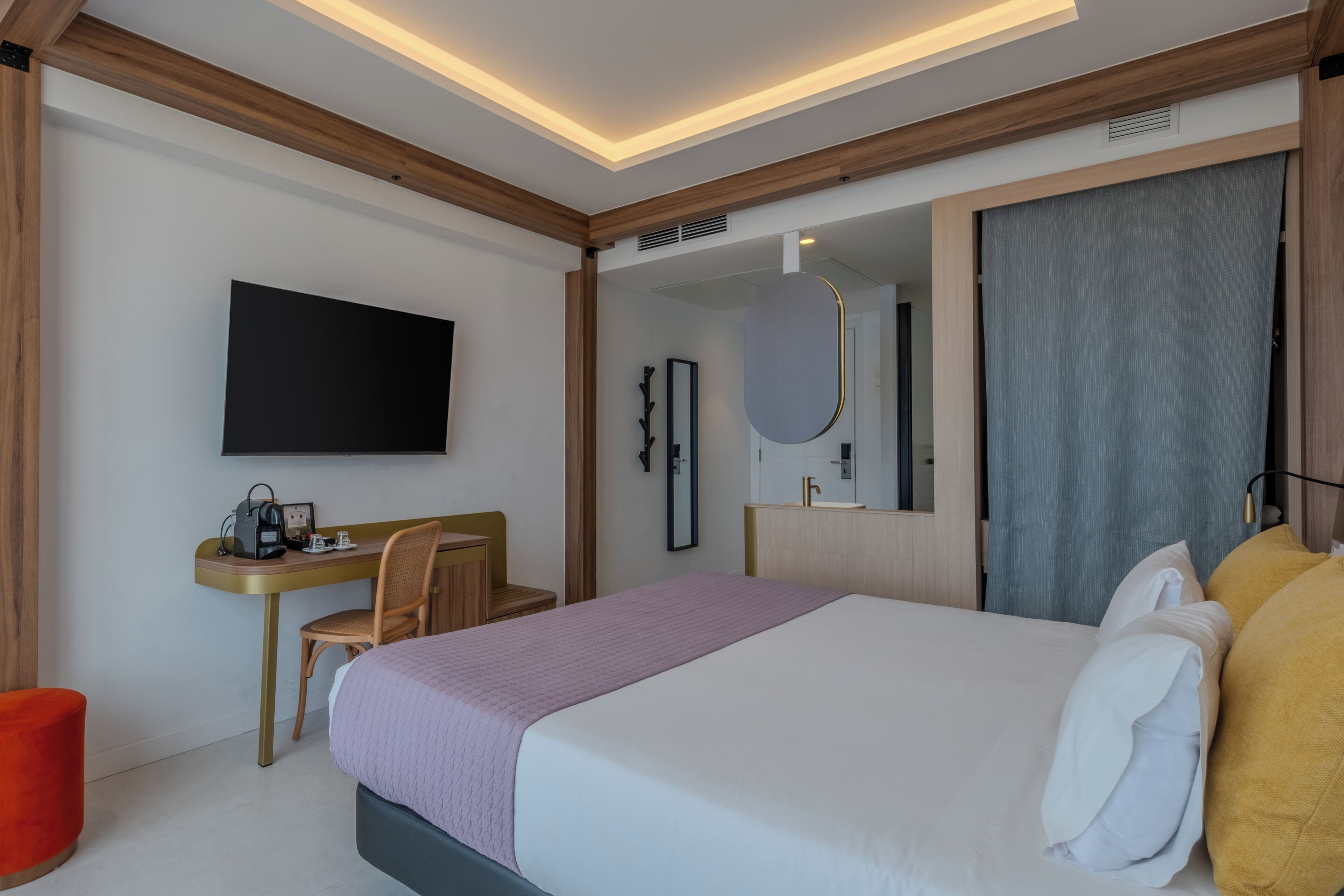 Ein modernes Hotelzimmer mit Bett, Schreibtisch, Sessel, Fernseher und einem großen Spiegel.