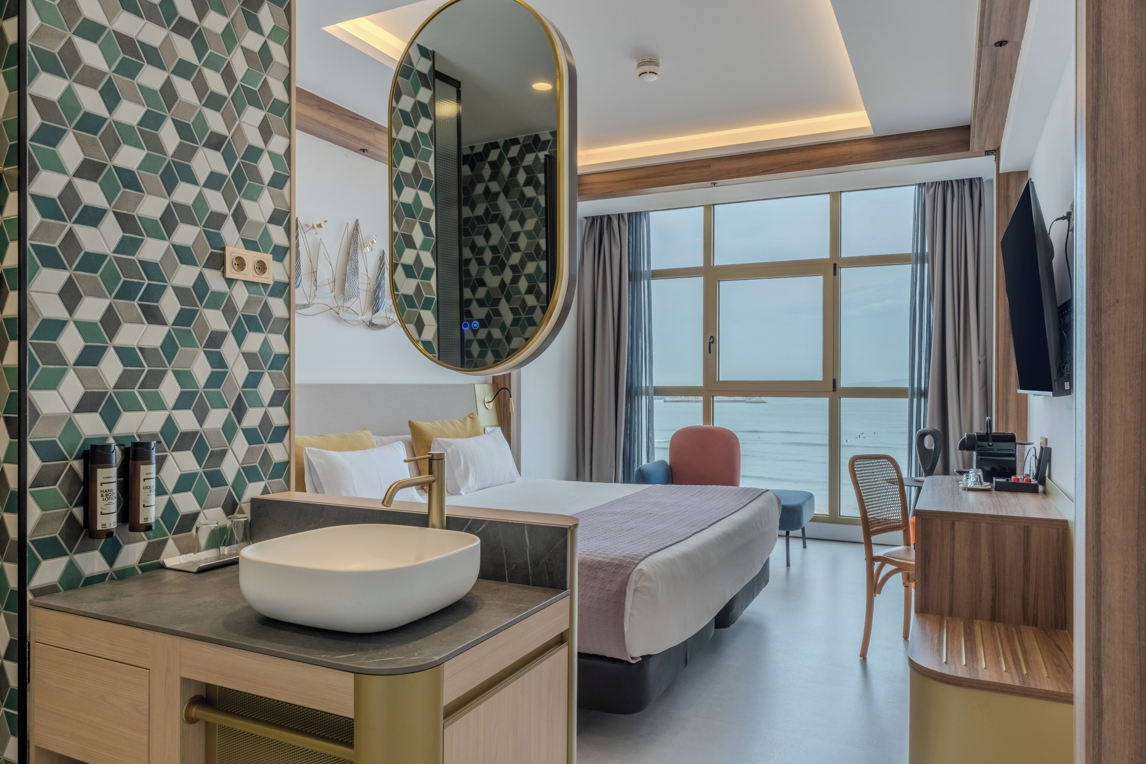 Habitación de hotel con una cama grande, un espejo redondo, un lavamanos, un escritorio y vista al mar.