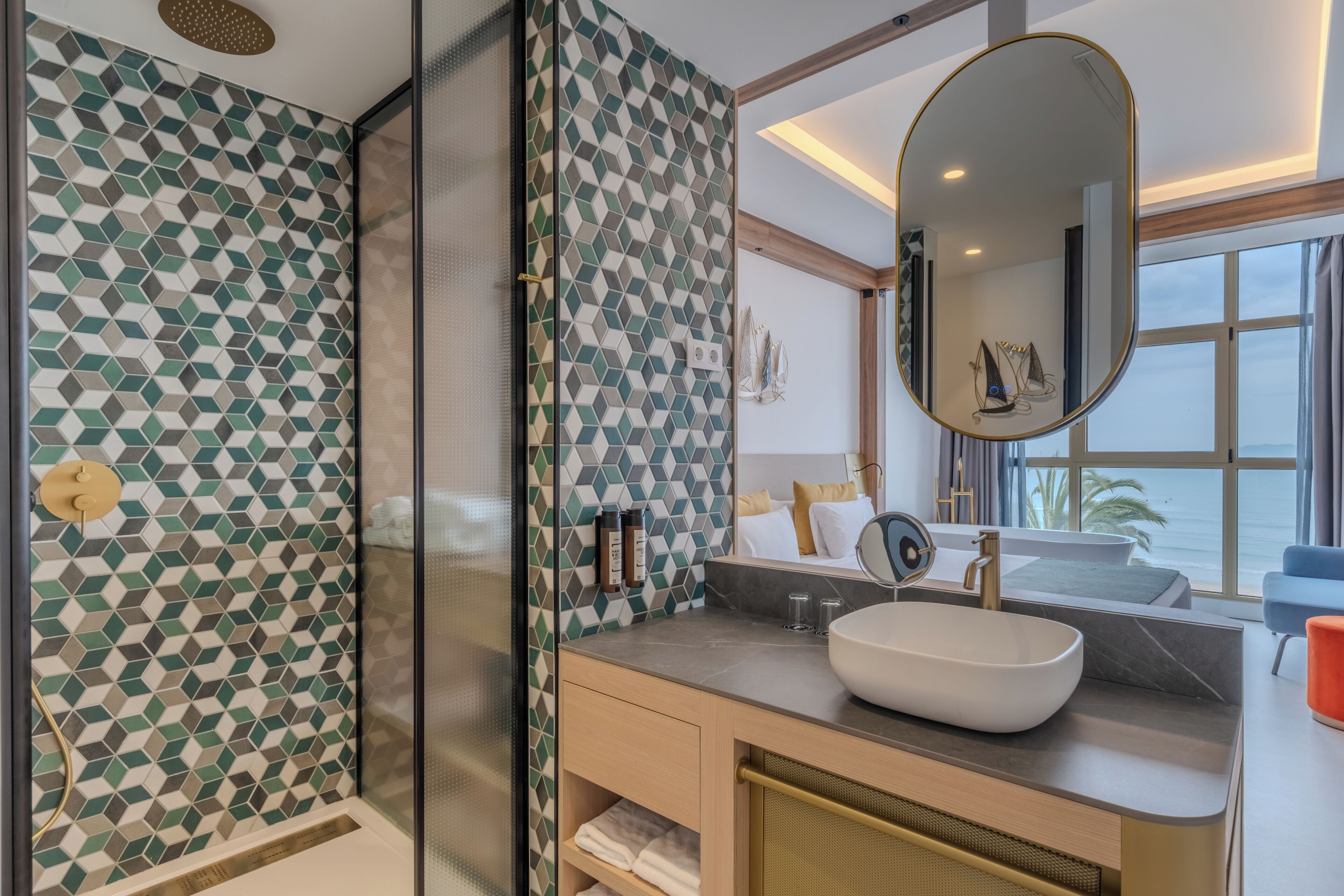 A modern bathroom with a geometric tile backsplash, a large oval mirror, a vessel sink, and a view of the sea through a large window.