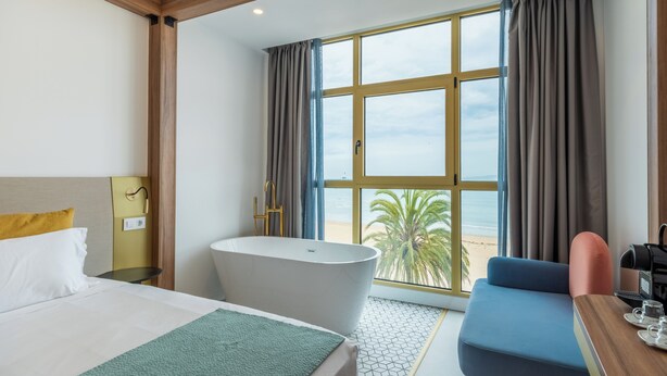 A hotel room with a bed, a bathtub, a desk, a chair, and a large window with a view of the beach. - Hotel Faranda Rias Altas, Ascend Hotel Collection (Oleiros)