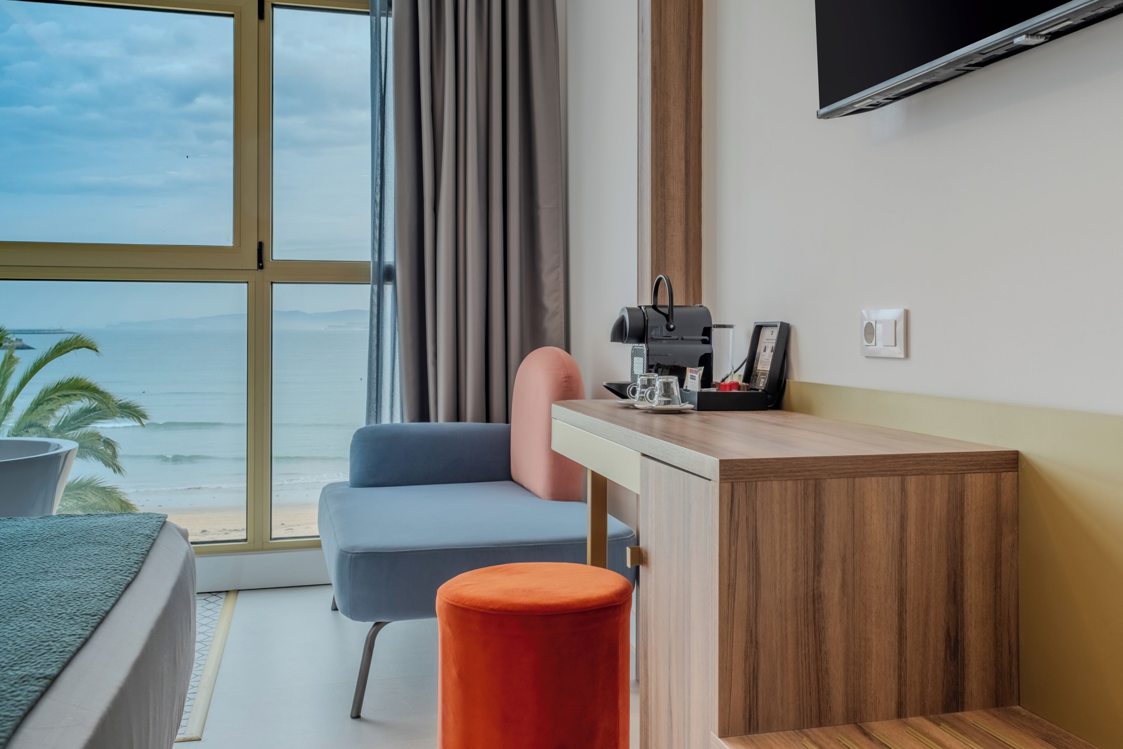A hotel room with a bed, a desk, a chair, a small orange stool, a television, and a view of the ocean.