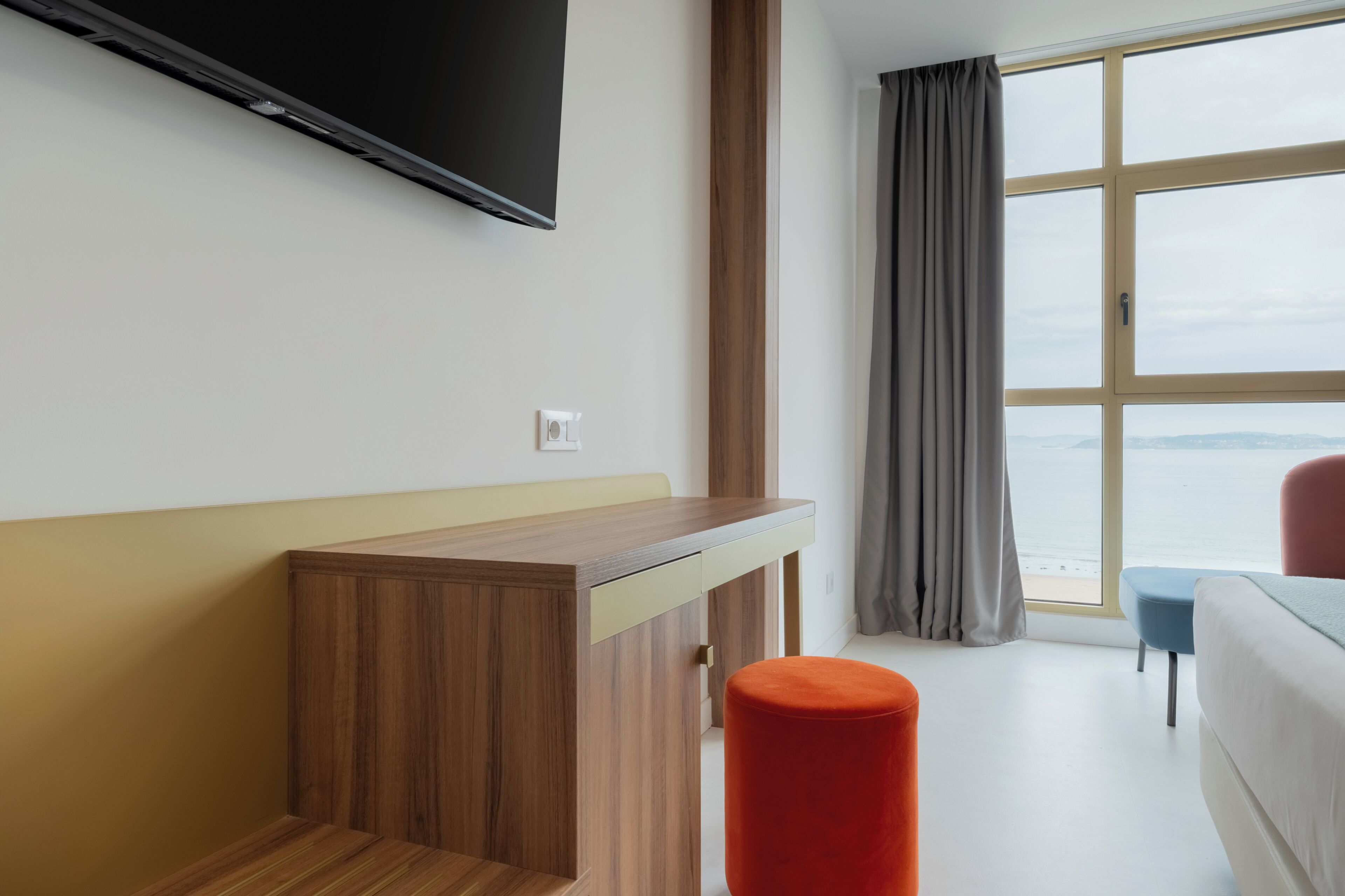 A hotel room with a wooden desk, a television, a window with a view of the sea, and a bed.