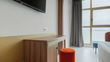 A hotel room with a wooden desk, a television, a window with a view of the sea, and a bed.