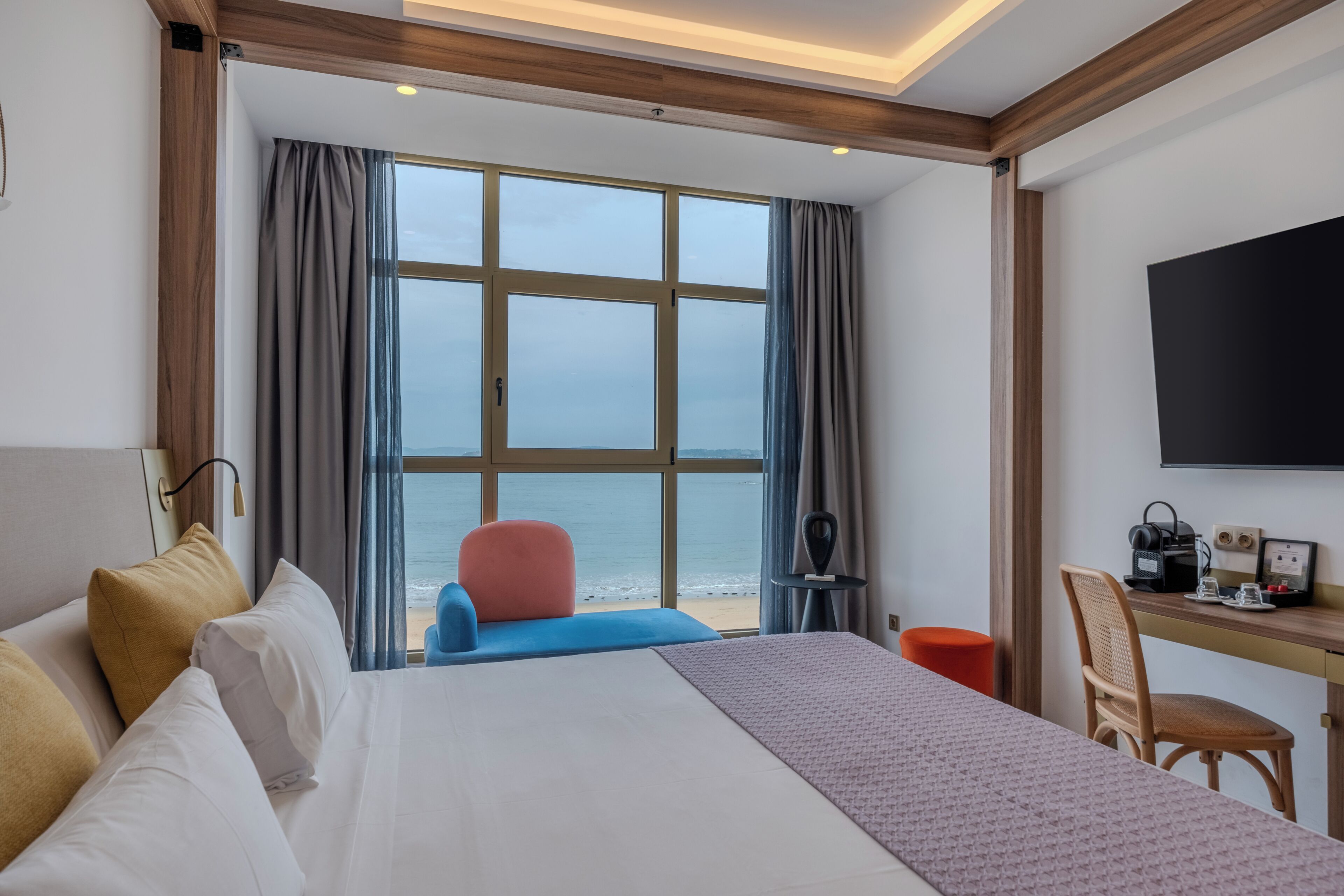 A hotel room with a bed, a desk with a chair, a TV, and a view of the sea through large windows.