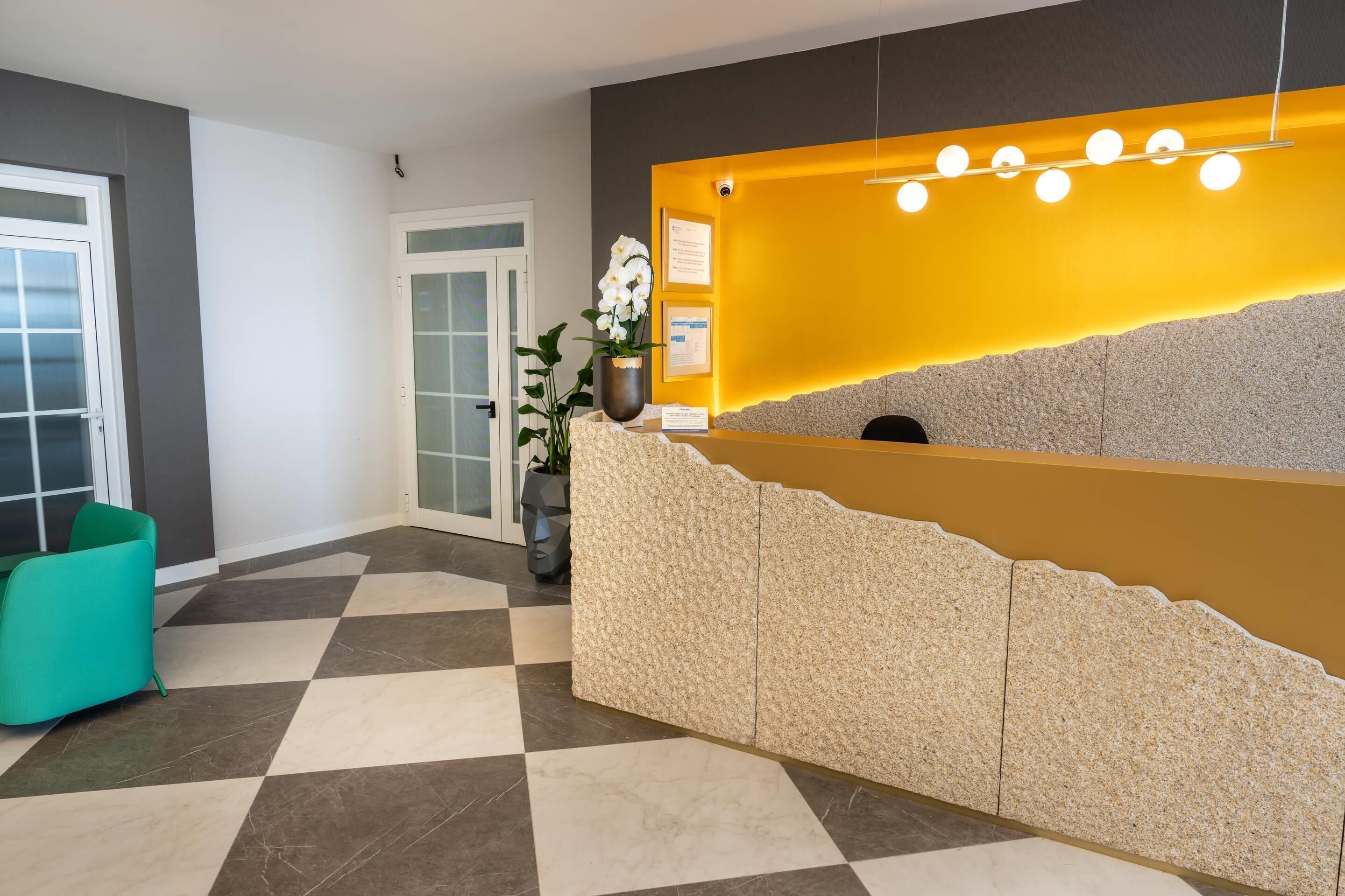 Un espace d’accueil avec un sol à carreaux, un comptoir de réception agrémenté d’un mur d’accent jaune, une plante en pot et un fauteuil de couleur turquoise.