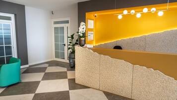 Un espace d’accueil avec un sol à carreaux, un comptoir de réception agrémenté d’un mur d’accent jaune, une plante en pot et un fauteuil de couleur turquoise.