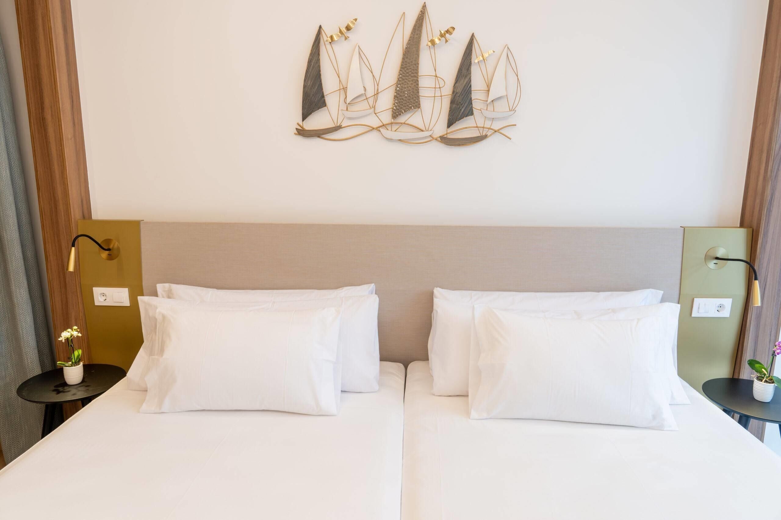Quarto de hotel com duas camas, uma obra de arte pendurada na parede retratando veleiros, uma mesa de cabeceira com abajur e uma pequena planta em vaso.