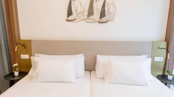 Quarto de hotel com duas camas, uma obra de arte pendurada na parede retratando veleiros, uma mesa de cabeceira com abajur e uma pequena planta em vaso.