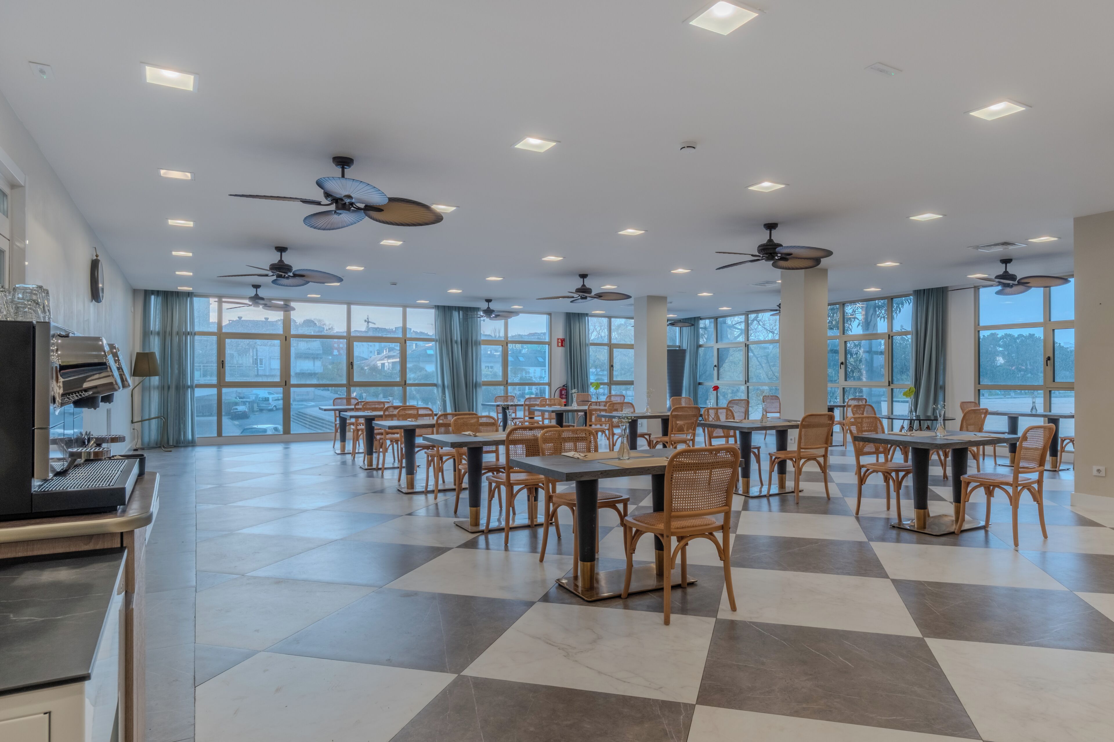 A spacious dining area with multiple tables and chairs, ceiling fans, and large windows offering a view of the cityscape.