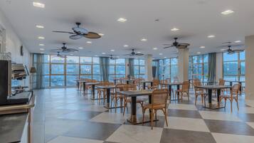 A spacious dining area with multiple tables and chairs, ceiling fans, and large windows offering a view of the cityscape.