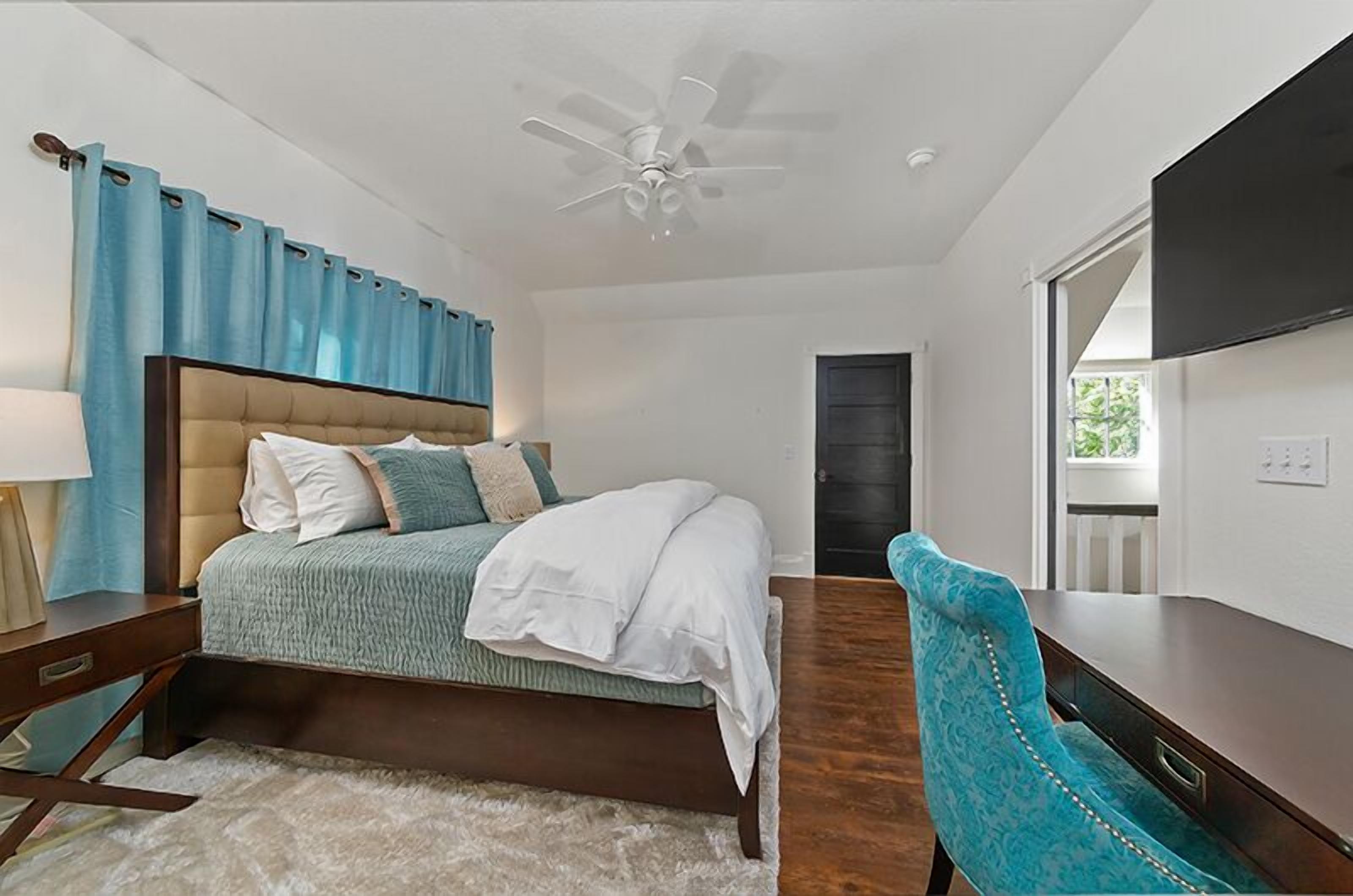A bedroom with a bed, a desk, a chair, a ceiling fan, and a door.