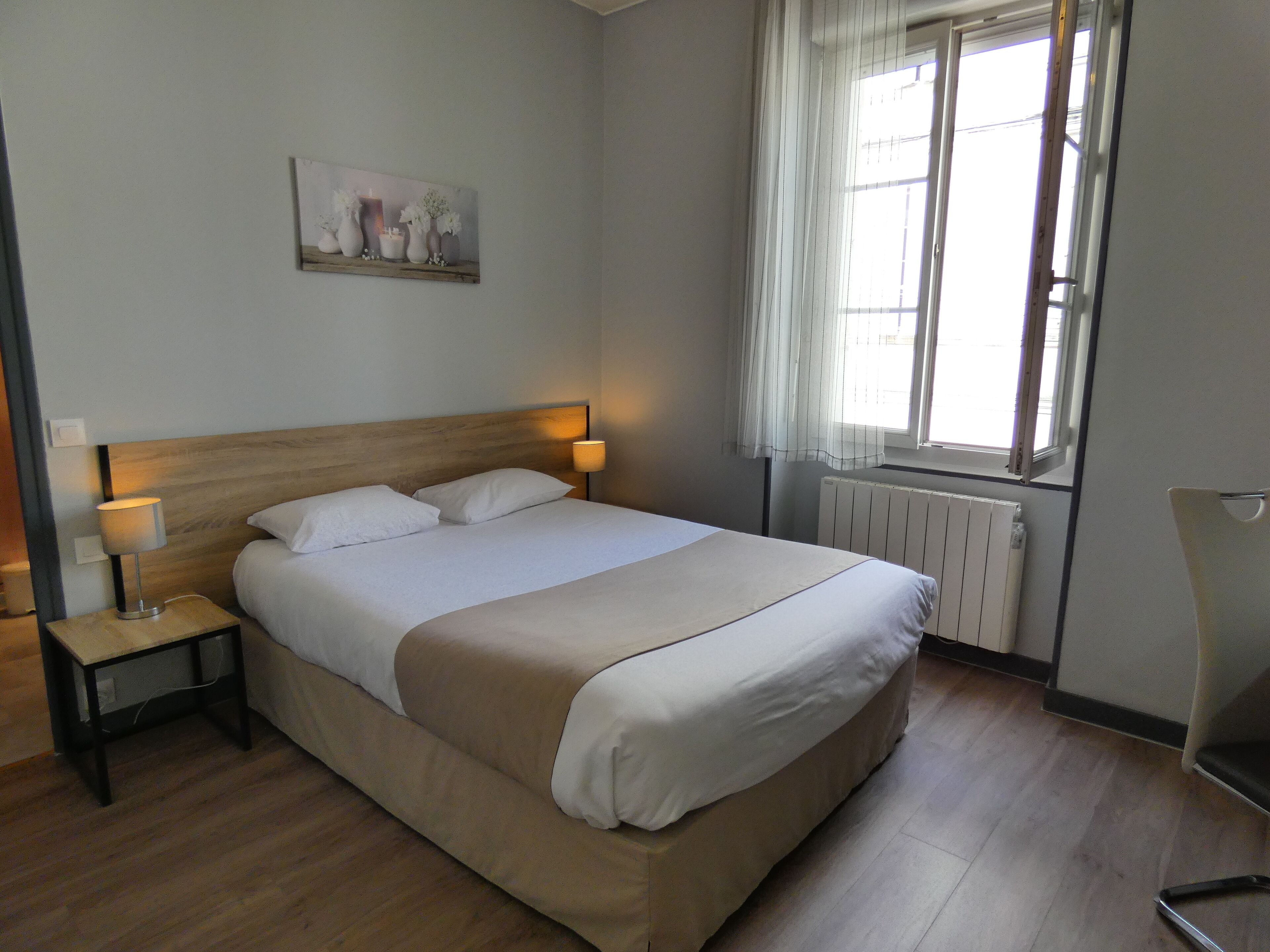 A neatly made bed with a wooden headboard, two bedside lamps, a radiator, and a window with curtains.