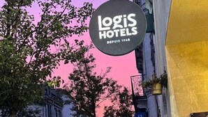 Blick auf eine Straße mit einem Gebäude, auf dem "Logis Hotels" steht, vor einem rosafarbenen Himmel.