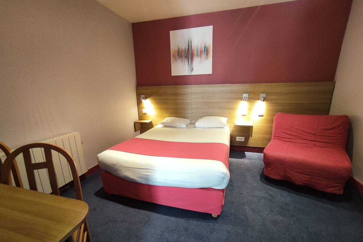 Ett hotellrum med en säng, två sängbord med lampor, en röd stol och ett träbord.