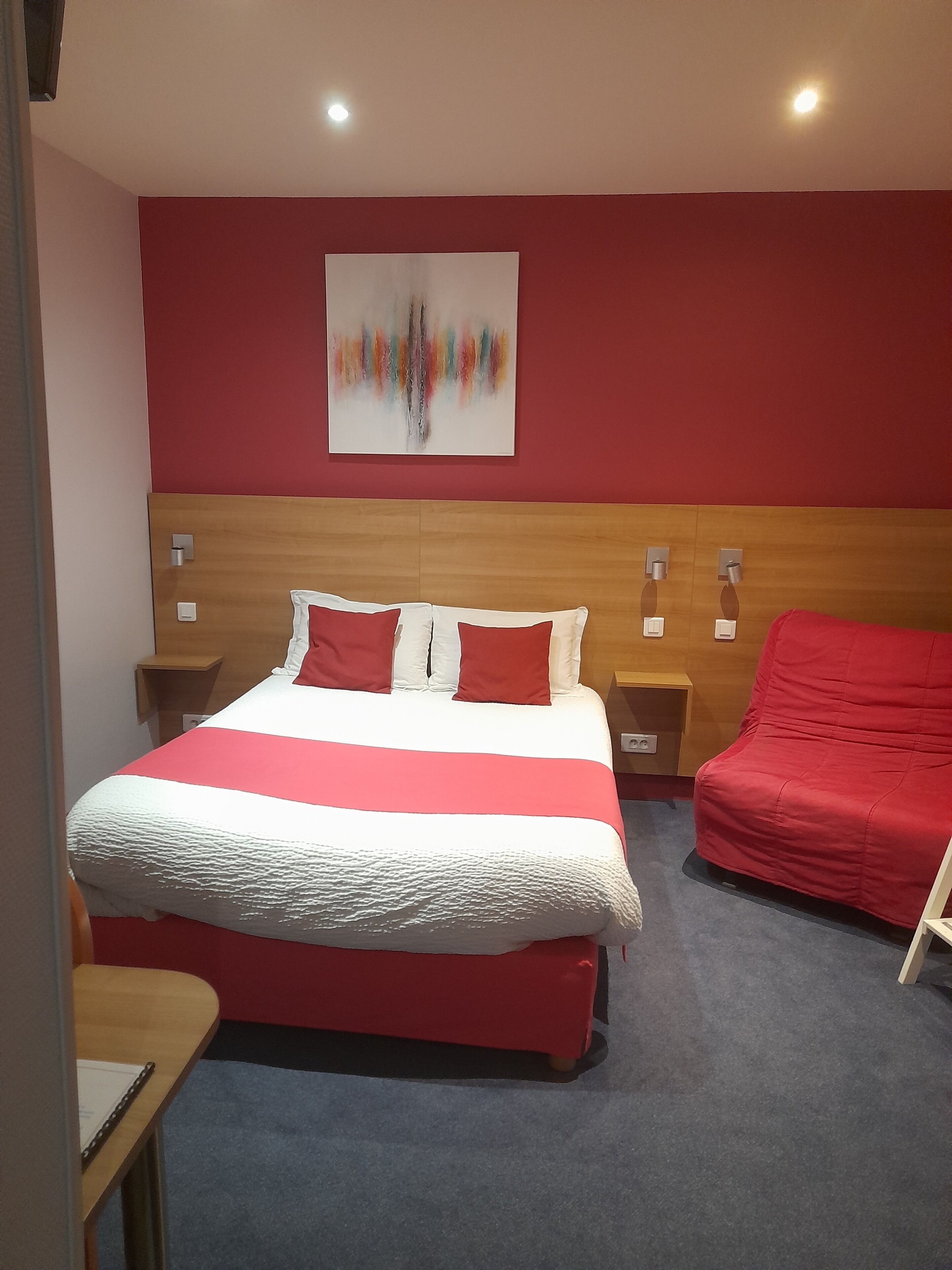 A hotel room with a large bed, a red chair, a wooden headboard, and a colorful abstract painting on the wall.