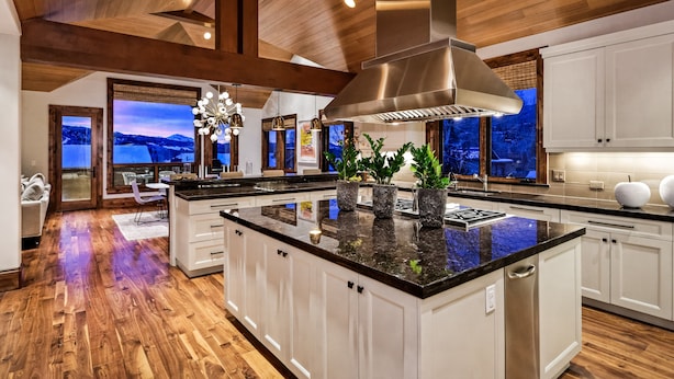 A modern kitchen with wooden floors, white cabinets, a large island, and a view of a mountainous landscape through the windows.