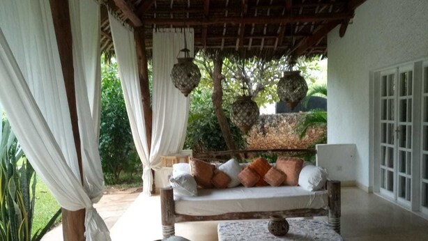 An outdoor seating area with a wooden bench and cushions, surrounded by potted plants and a thatched roof. - Diamond home 001 (Malindi)