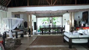 A spacious indoor area with a high ceiling, a central seating area with sofas and a dining table, and a view of a tropical outdoor space. - Diamond home 001 (Malindi)