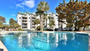 A clear swimming pool with lounge chairs and a multi-story building in the background.