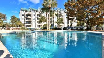 A clear swimming pool with lounge chairs and a multi-story building in the background.