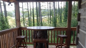 A wooden deck with a round table and two chairs, surrounded by a wooden railing, overlooking a forested area.