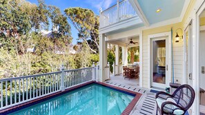 A pool with a white deck, a covered patio with outdoor furniture, and a view of trees and a house.