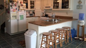Una cocina con armarios blancos, una isla central con taburetes, un refrigerador, un microondas y un dispensador de agua.