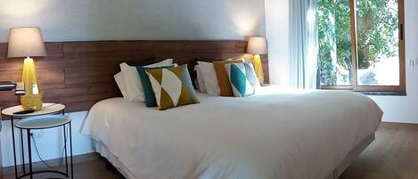 Un dormitorio con una cama grande, múltiples almohadas, lámparas de noche y una ventana con vistas a zonas verdes.