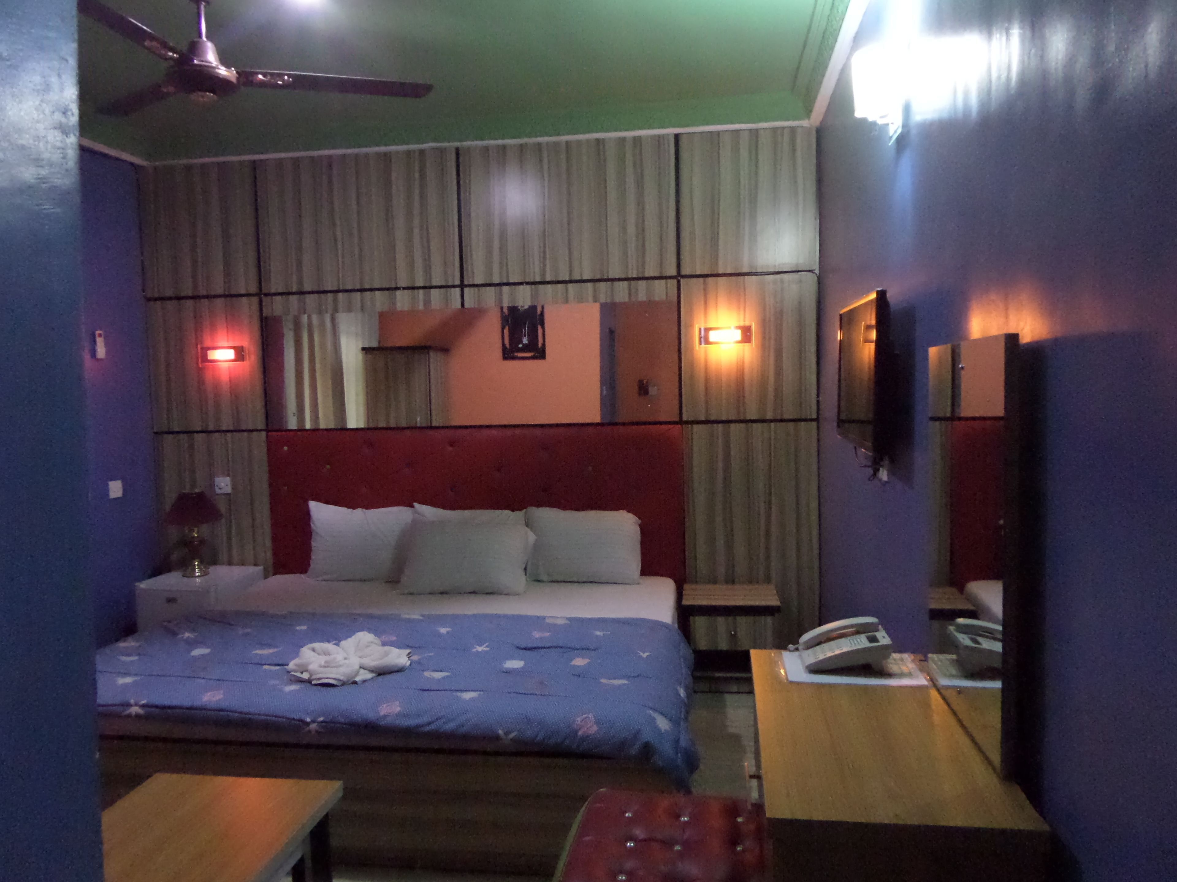 Habitación de hotel con cama, escritorio, silla, ventilador de techo y televisor montado en la pared.