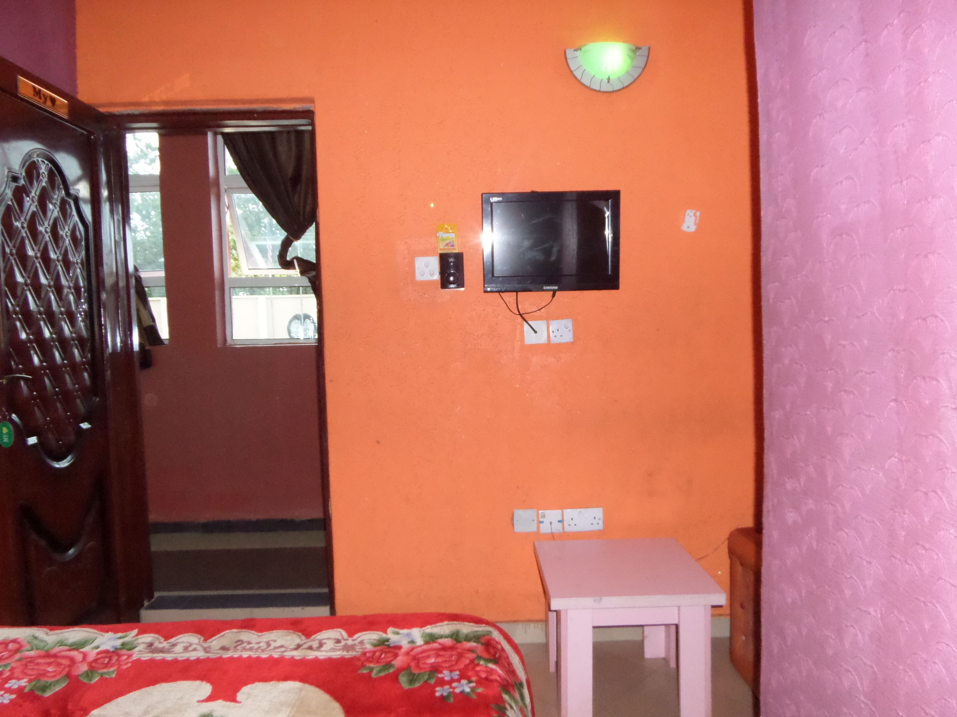 Una habitación con pared naranja, televisor montado en la pared, cama con colcha floral, una mesita y una puerta con manija.