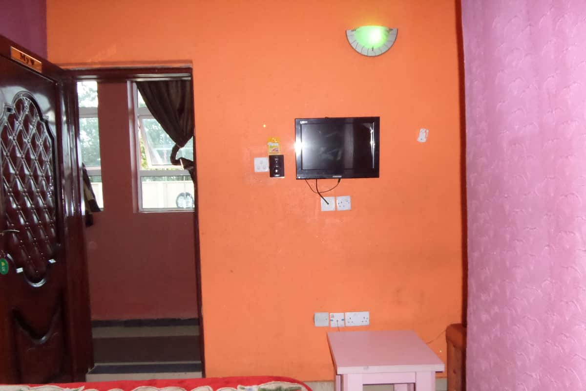 Een kamer met een oranje wand, een aan de muur bevestigde televisie, een bed met een bloemenprint en een klein tafeltje, plus een deur met een handgreep.