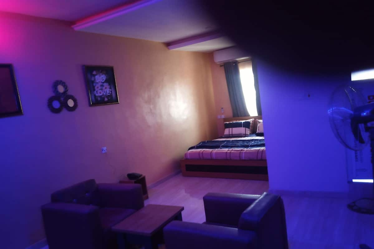 Een kamer met een bed, een bureau en een stoel. Er is een raam met gordijnen en een ventilator aan de muur bevestigd.