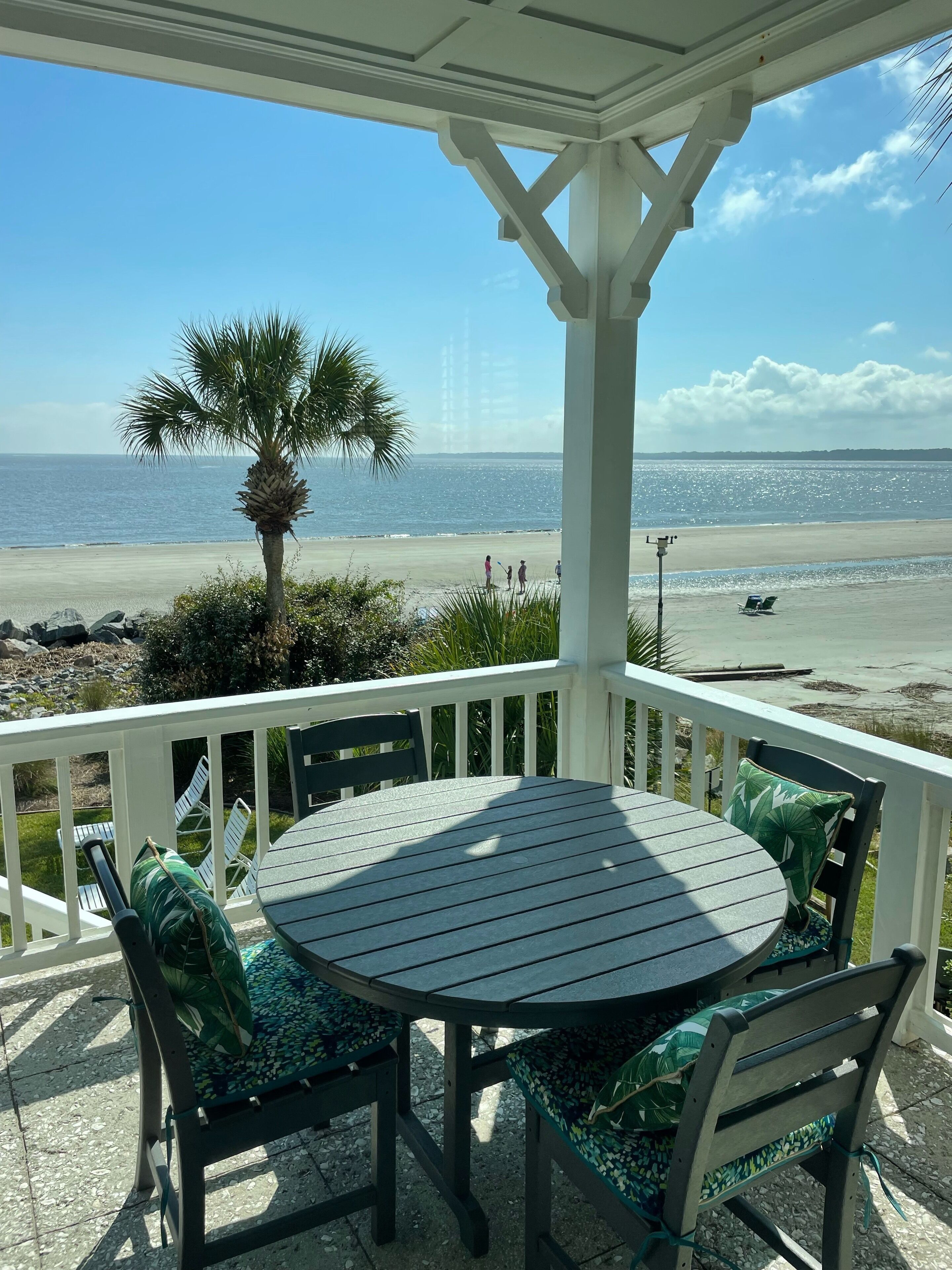 Une terrasse couverte avec une table ronde et des chaises, donnant sur une plage avec un palmier et une vue sur l’océan.