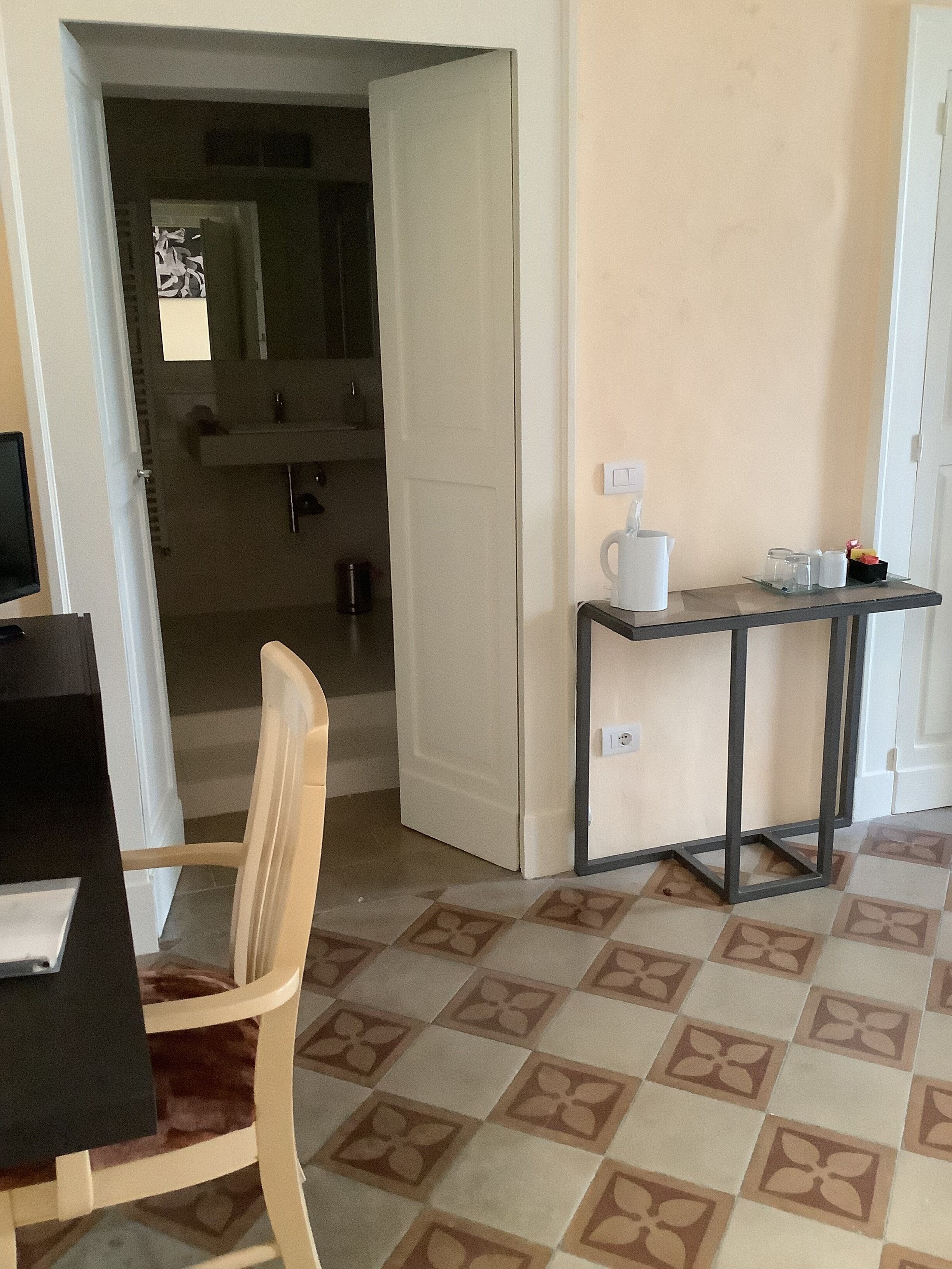 A room with a dining table, a chair, a small table with a coffee maker, and a tiled floor.