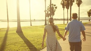 Ein Paar geht Hand in Hand entlang einer Uferpromenade mit Palmen und Wasser im Hintergrund.
