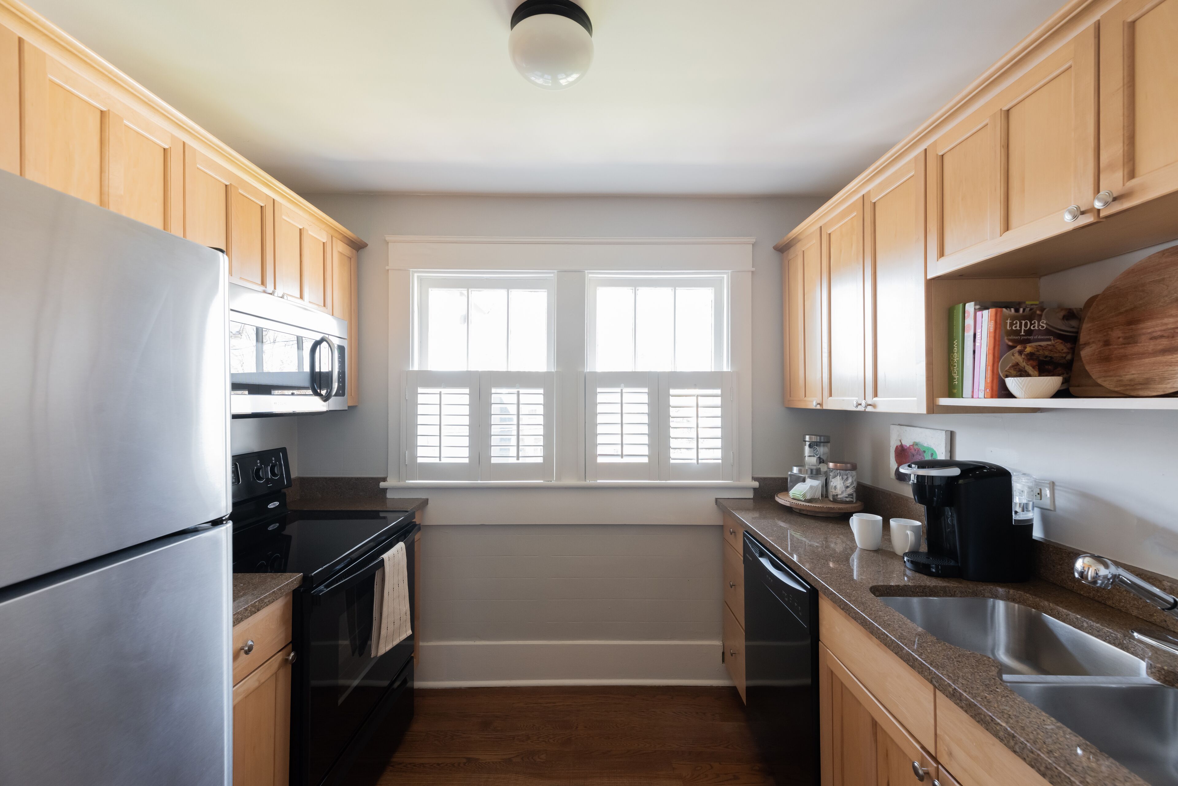 A kitchen with wooden cabinets, a stainless steel refrigerator, a microwave, an oven, a dishwasher, a coffee maker, and a window with blinds.