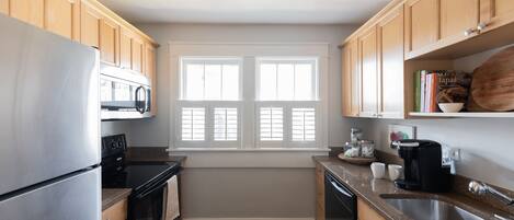 A kitchen with wooden cabinets, a stainless steel refrigerator, a microwave, an oven, a dishwasher, a coffee maker, and a window with blinds.