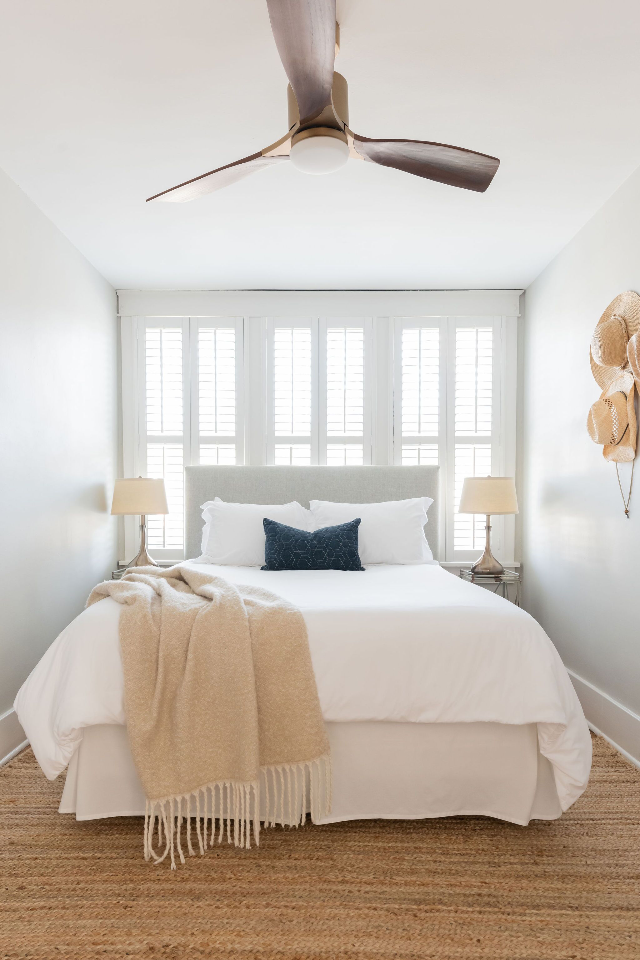 Une chambre avec un grand lit, deux tables de chevet avec des lampes, un ventilateur de plafond et une fenêtre avec des stores.