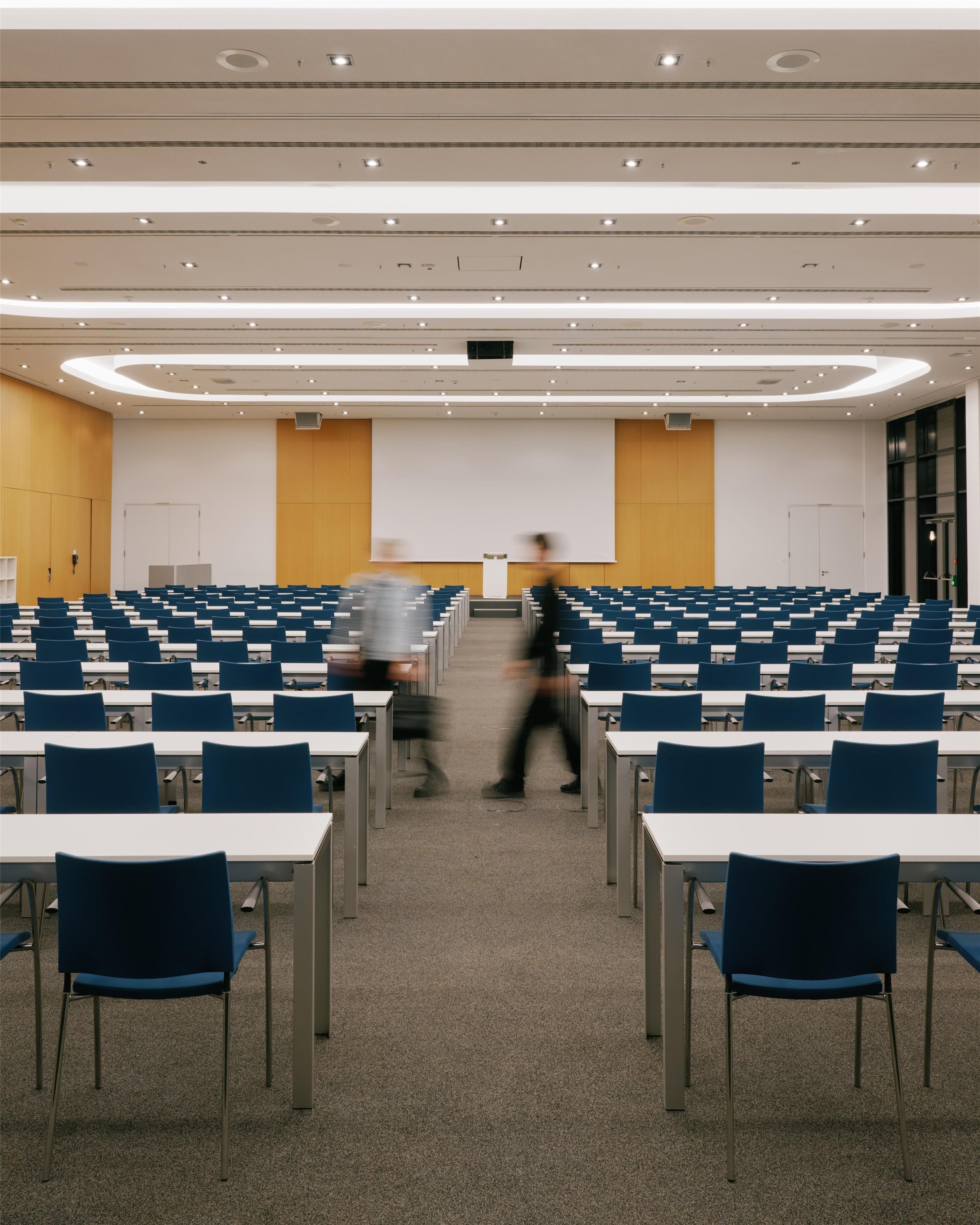 A conference room with rows of tables and chairs, a large screen at the front, and a person walking in the aisle.