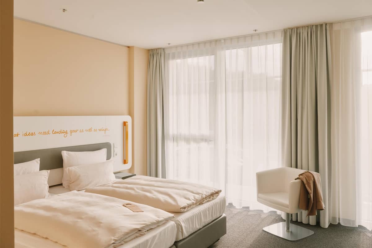 Une chambre d’hôtel avec un lit, une chaise et une fenêtre avec des rideaux légers.