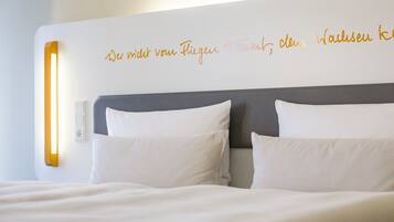 A hotel room with a bed, two pillows, a bedside table, and a wall with a light fixture and text.