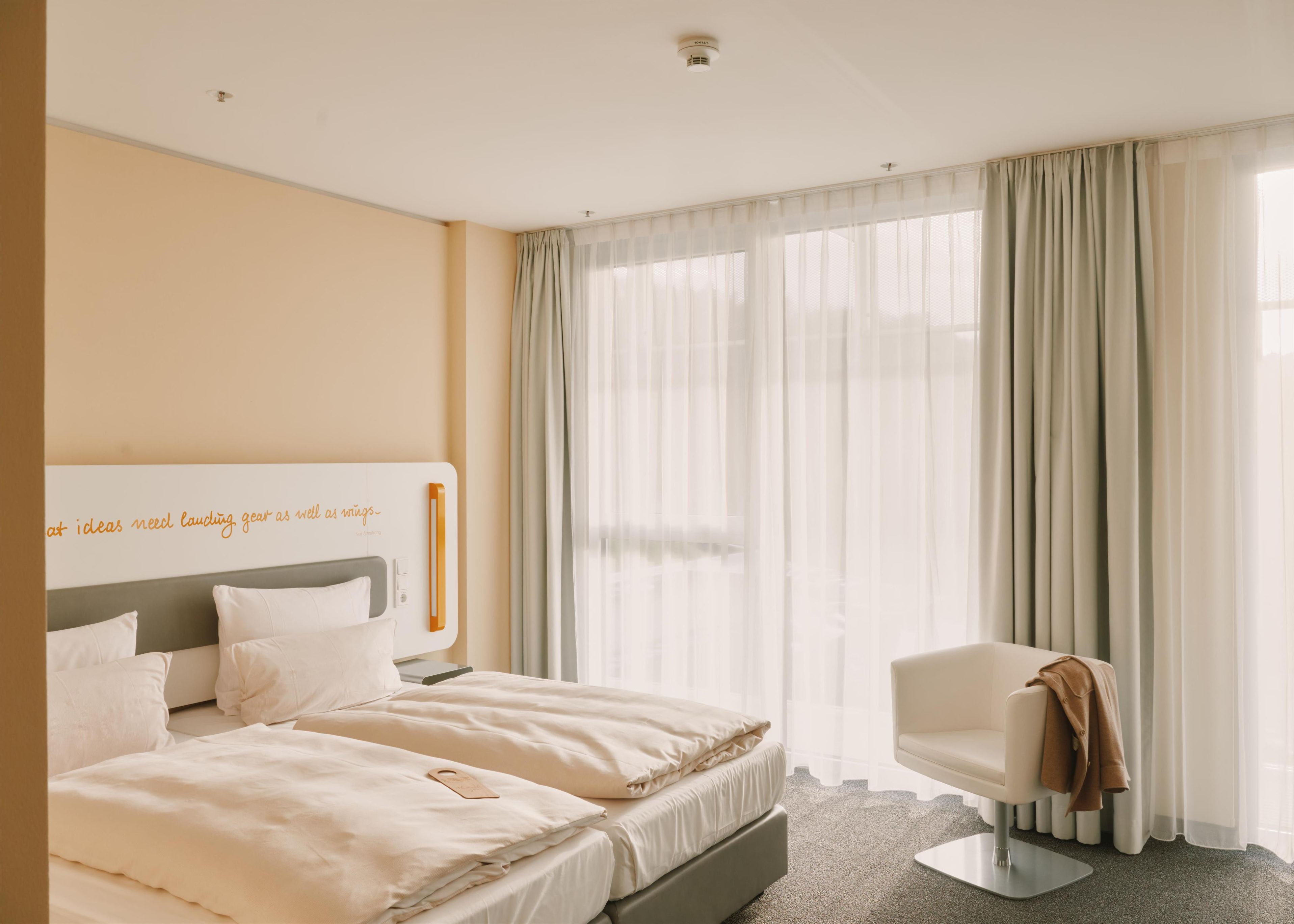 Ein Hotelzimmer mit einem Bett, einem Sessel und einem Fenster mit durchscheinenden Vorhängen.