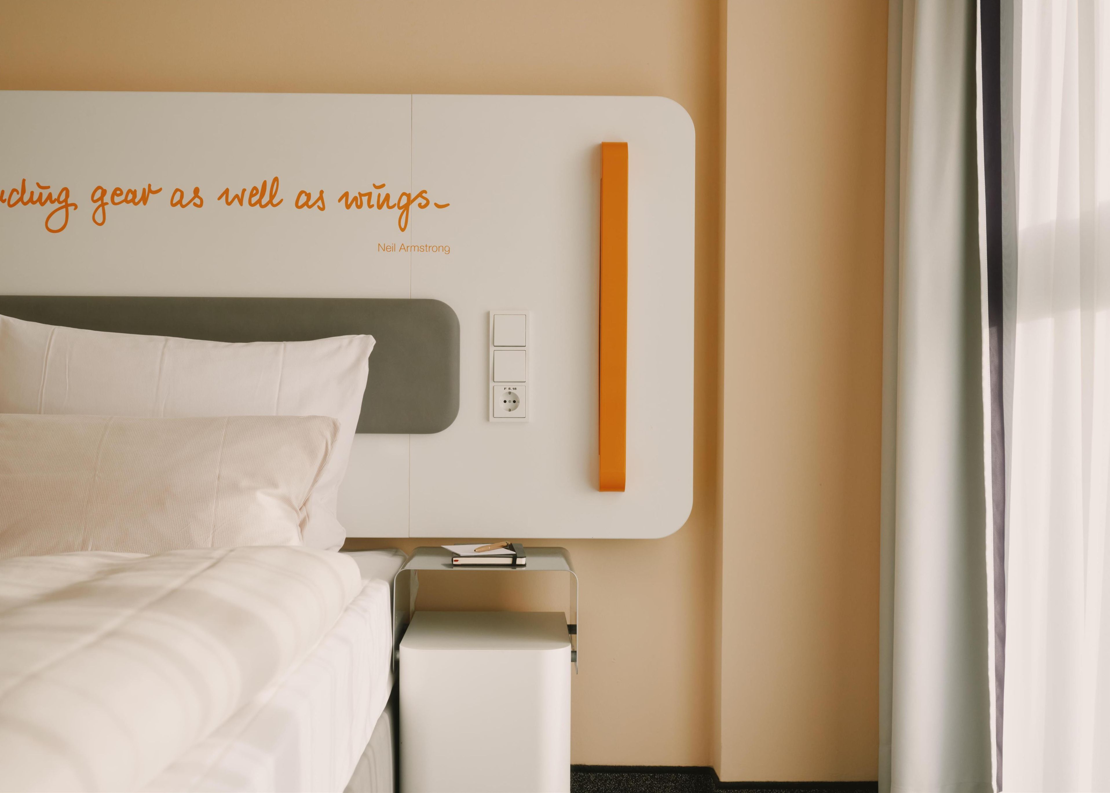 A modern hotel room with a neatly made bed, a bedside table with a book, and a wall-mounted orange strip.