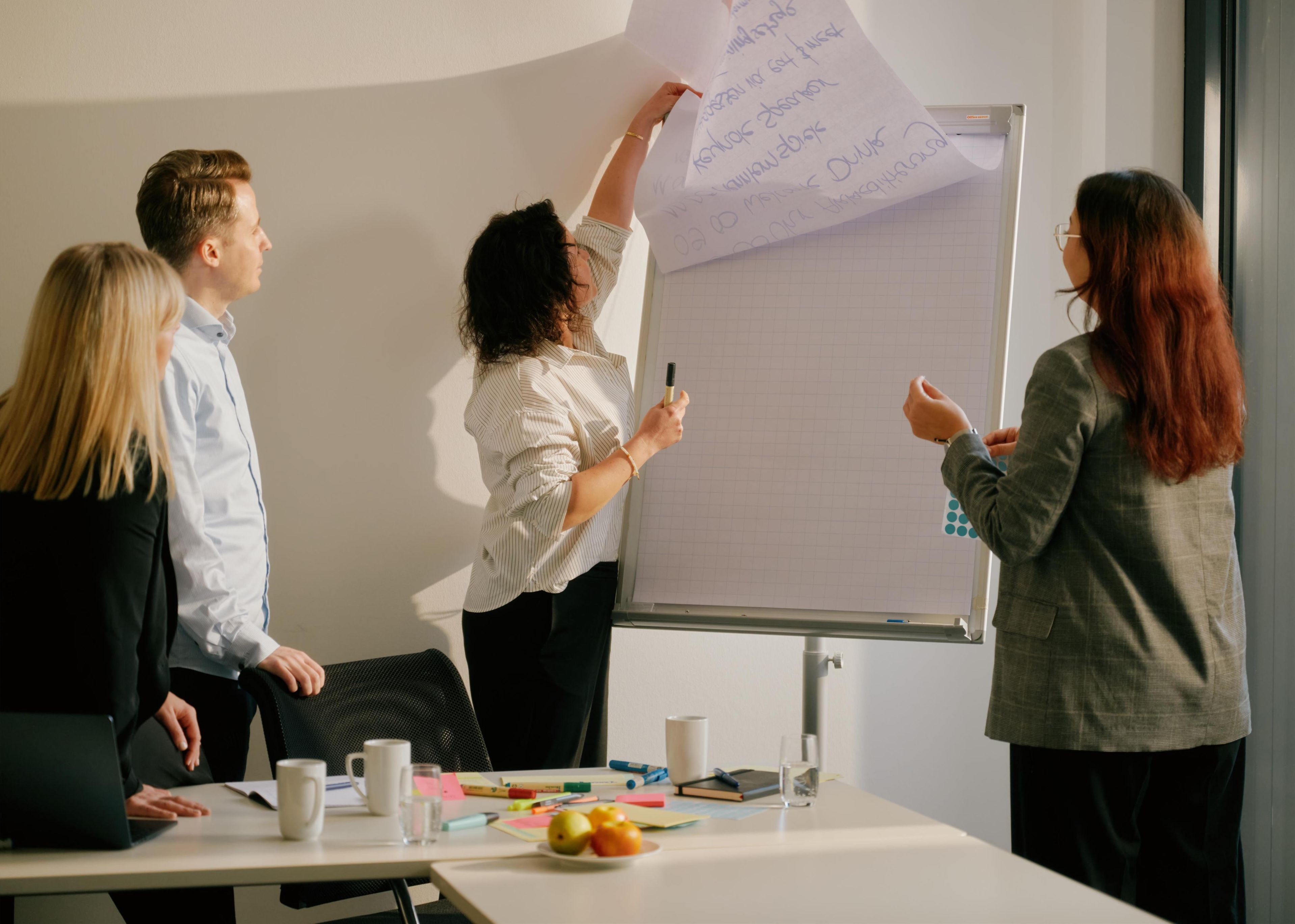 A group of people in a meeting room with a whiteboard, a laptop, and some stationery items on the table.