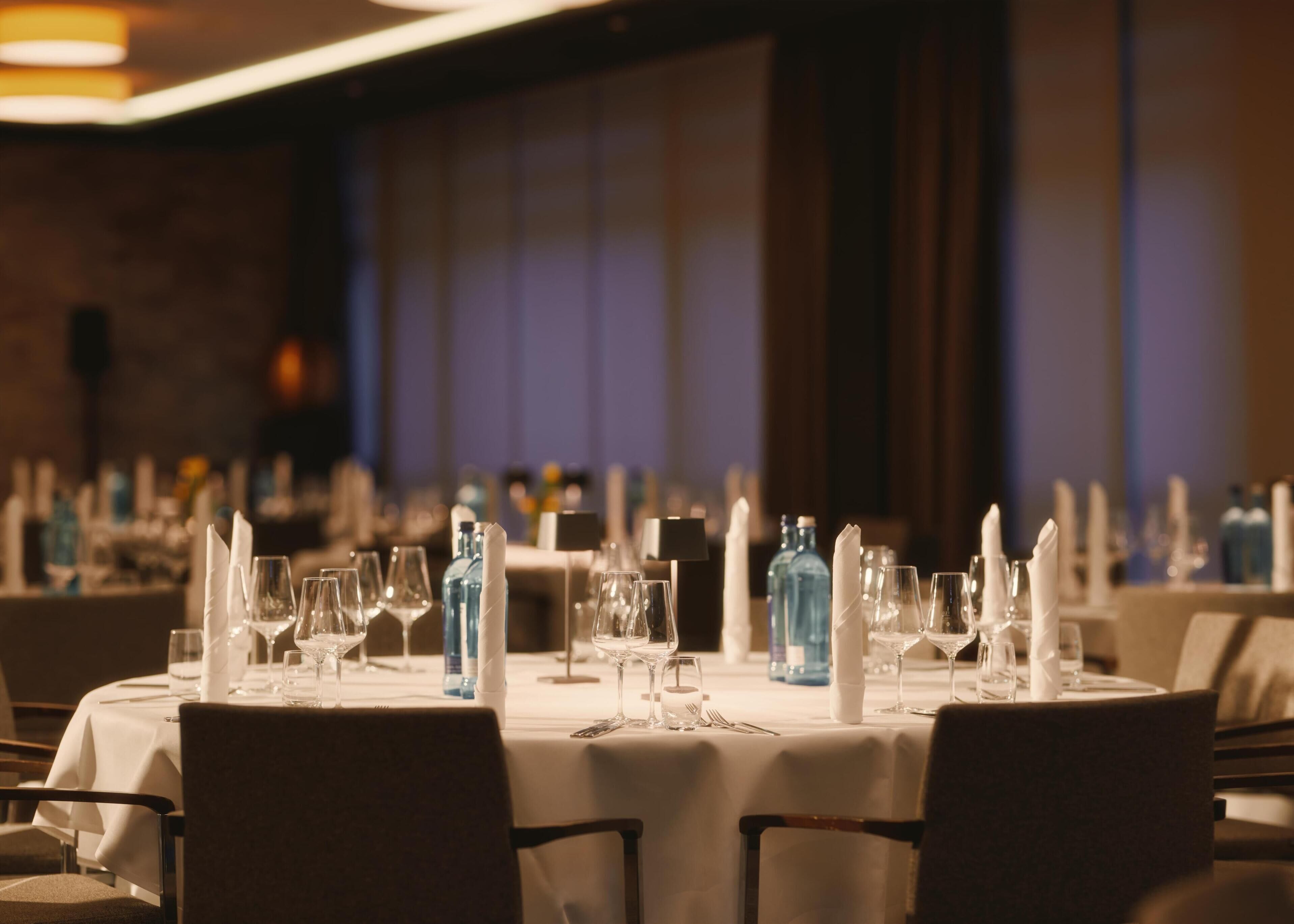 A restaurant with round tables set for dining, featuring white tablecloths and neatly arranged glassware.