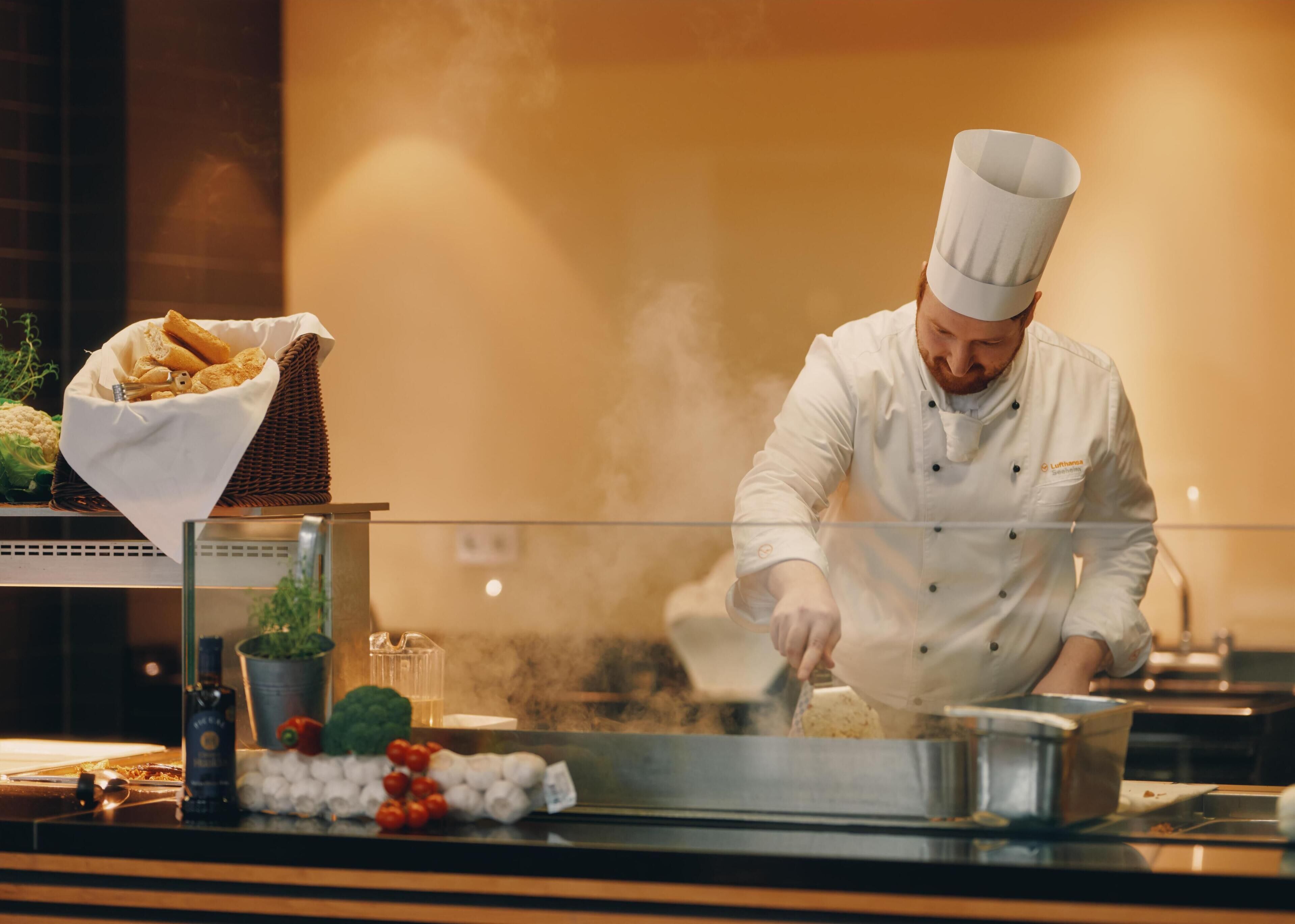 A chef in a white uniform with a chef's hat, preparing food in a kitchen with a steam vent. Visible items include a basket of bread, a bottle of olive oil, and a bowl of tomatoes.