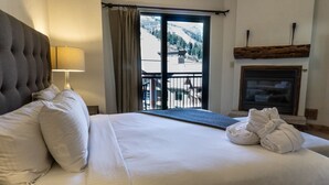 A hotel room with a large bed, a nightstand with a lamp, a view of mountains through a sliding door, and a fireplace. - Four Bedroom Penthouse in Canyons Village by RedAwning (Park City)