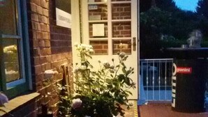 A brick building with a glass door, a small garden with roses, and a trash bin labeled "granini". - BASALT Hotel Restaurant Lounge (Hohwacht)