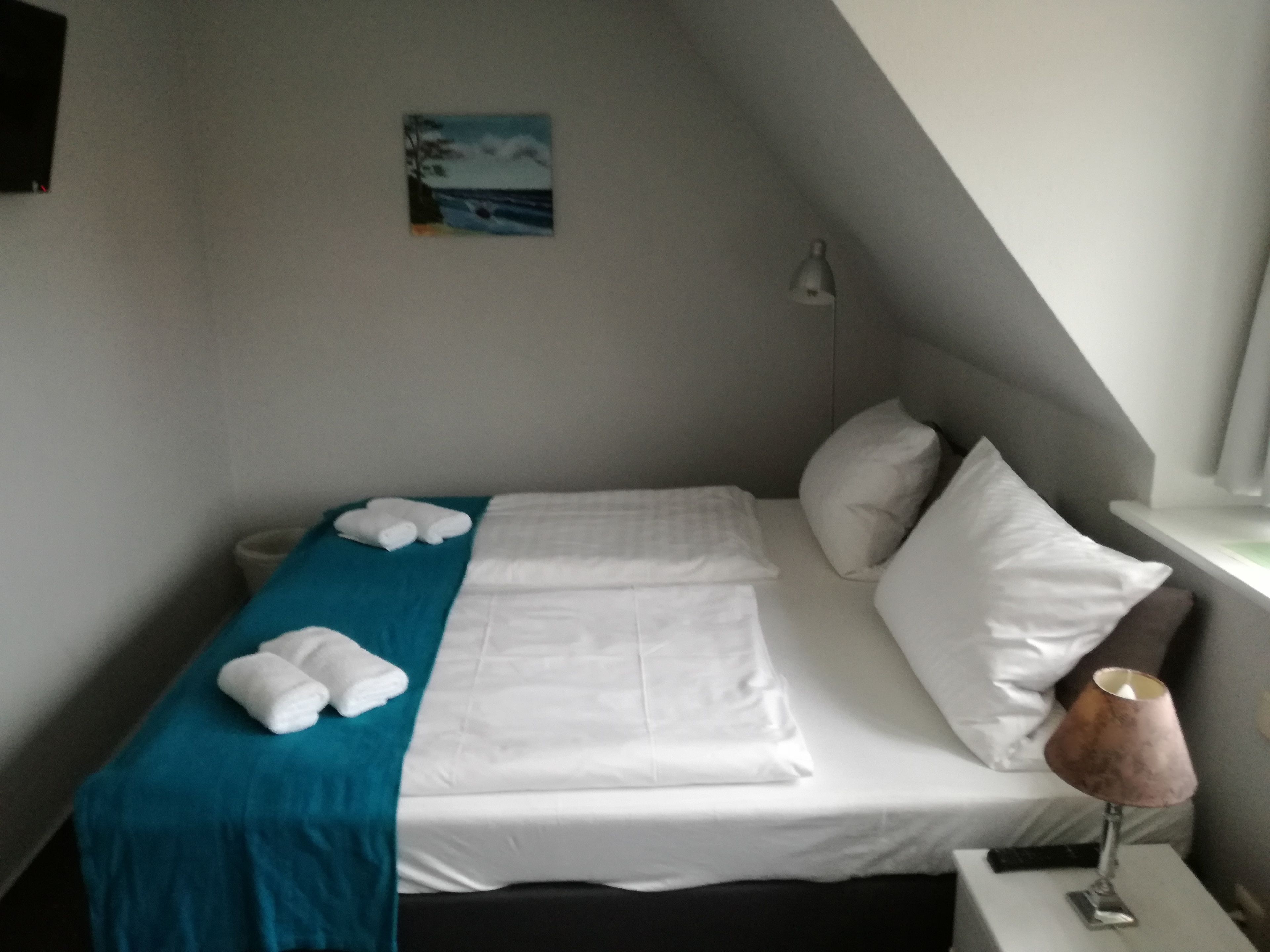 A neatly made bed with white linens, a teal blanket, and two rolled-up towels. There is a small painting on the wall above the bed, a bedside lamp, and a window with a view of the sky.